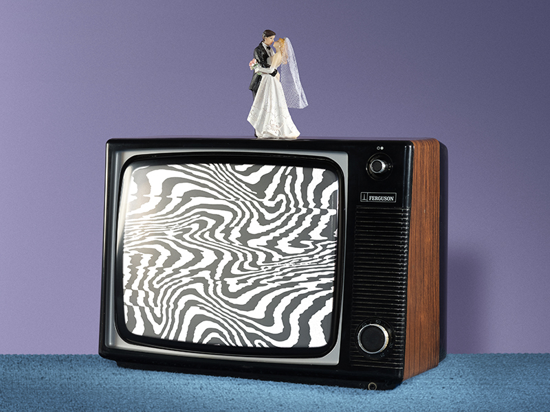 A vintage TV with static, wavy black-and-white lines on the screen sits on blue carpet. On top of the TV is a bride and groom wedding cake topper, set against a purple background.