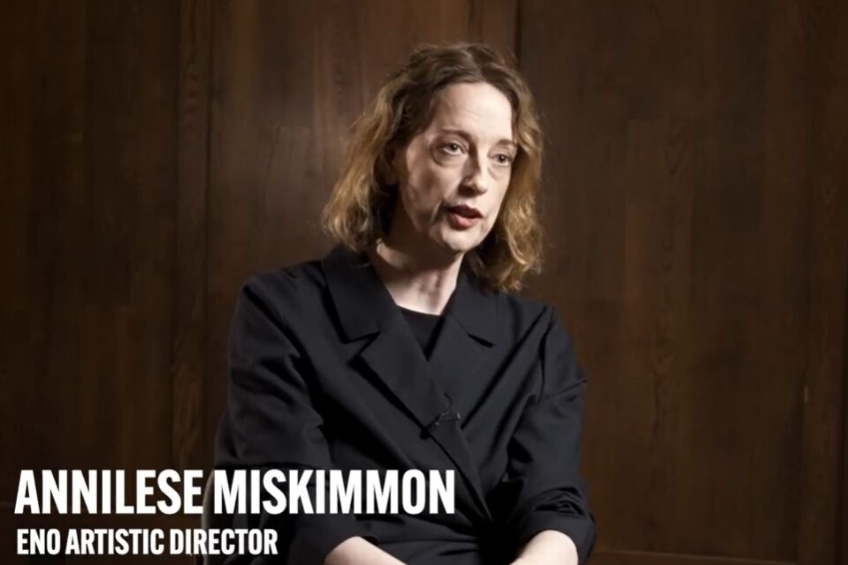 A woman with light skin and shoulder-length brown hair, wearing a dark blazer, sits for an interview in front of a wooden wall. Text reads Annilese Miskimmon, ENO Artistic Director with an ENO logo in the top right corner.