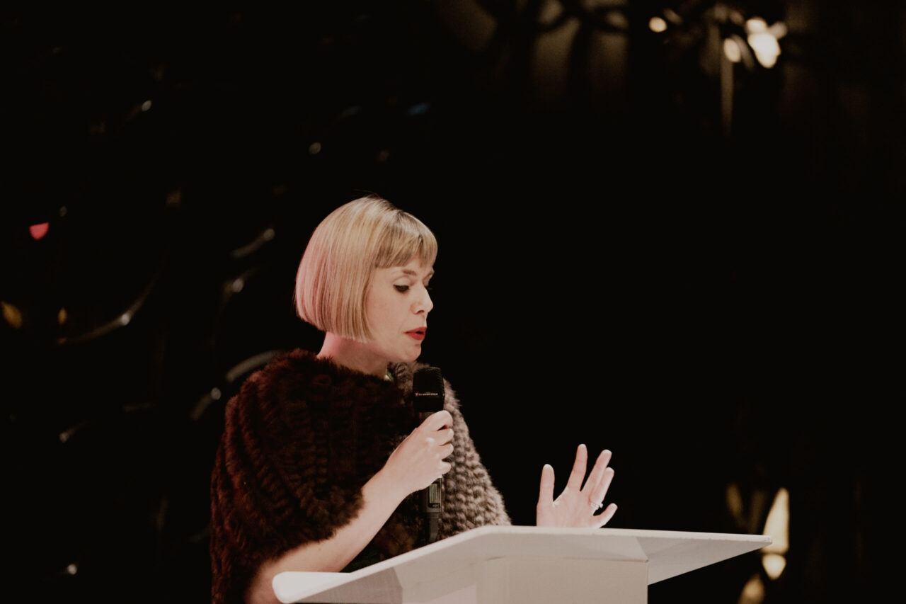 Jenny, a woman with short blonde hair, wearing a dark textured shawl, stands at a lectern holding a microphone and speaking with one hand raised, against a dark, out-of-focus background.