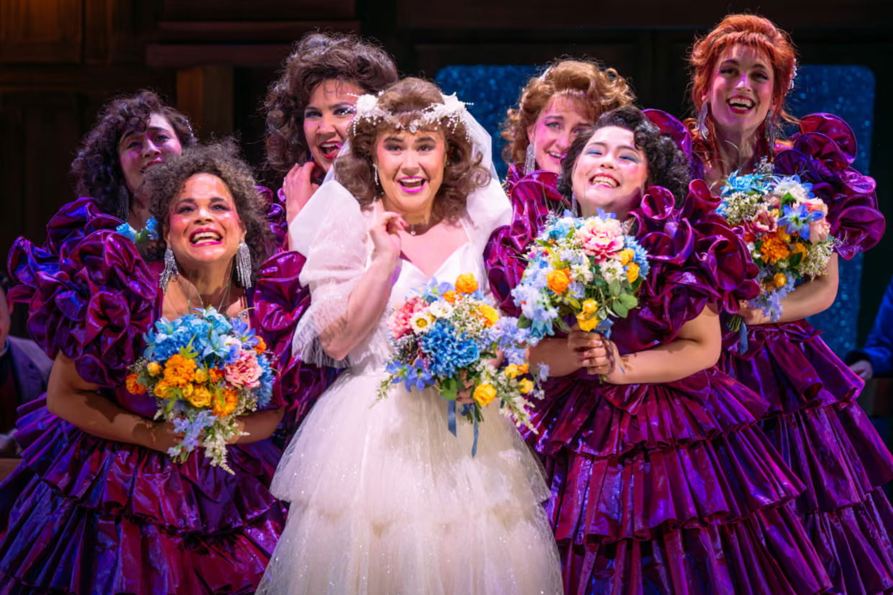 A smiling bride in a white wedding dress holds a bouquet and is surrounded by six bridesmaids in matching purple ruffled dresses, posing joyfully on stage.