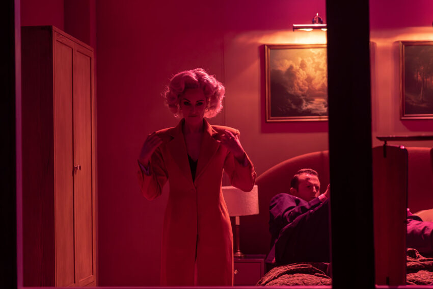 A woman in a red coat stands in a dimly lit room with pink lighting, adjusting her collar. A man lies on a bed in the background, propped up with pillows. Paintings and a lamp decorate the room.