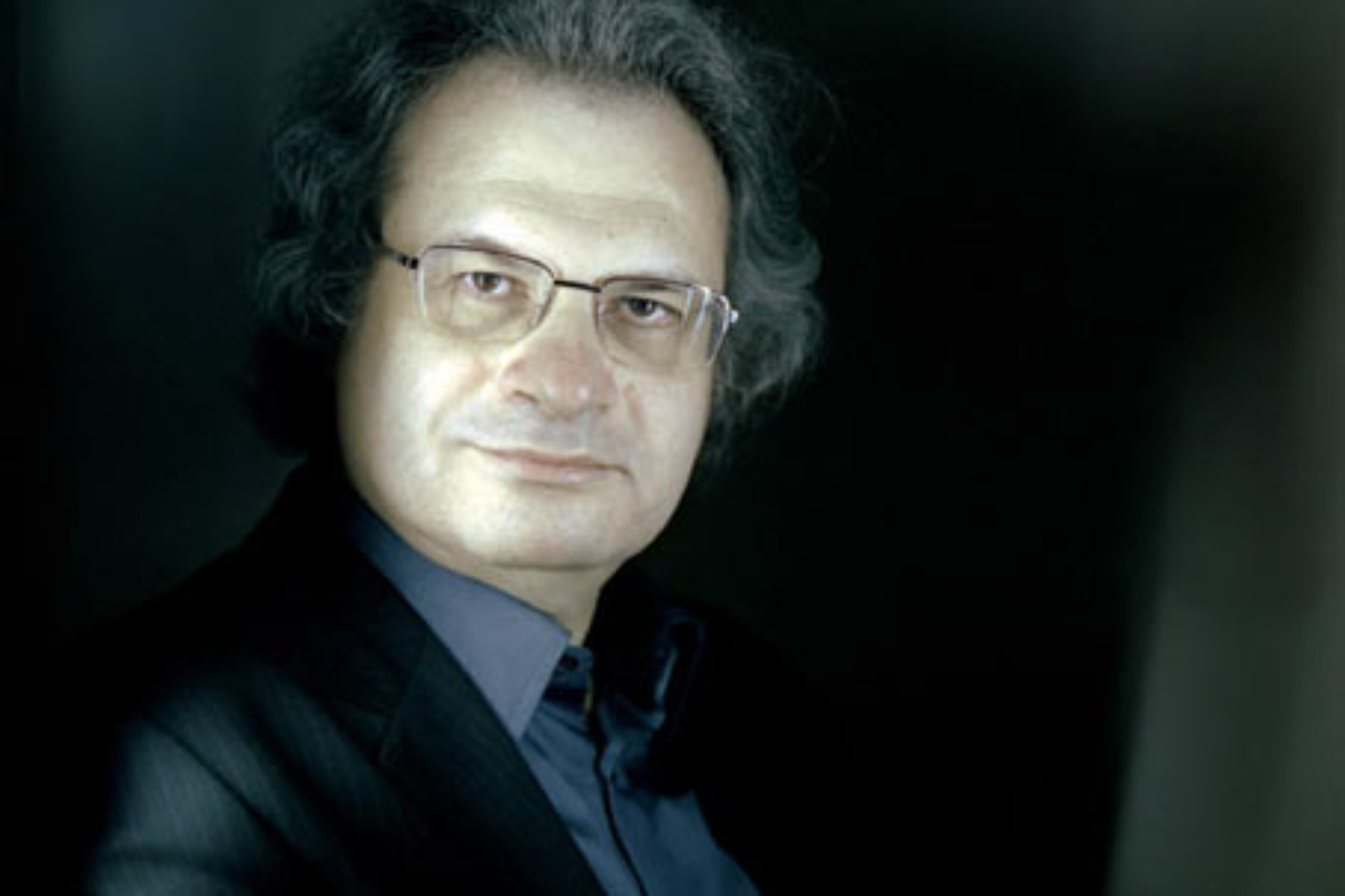 A middle-aged man with grey hair and glasses, in a dark suit and blue shirt, looks at the camera with a slight smile—an introduction reminiscent of an Adriana Mater performance, set against a dark, softly lit background.