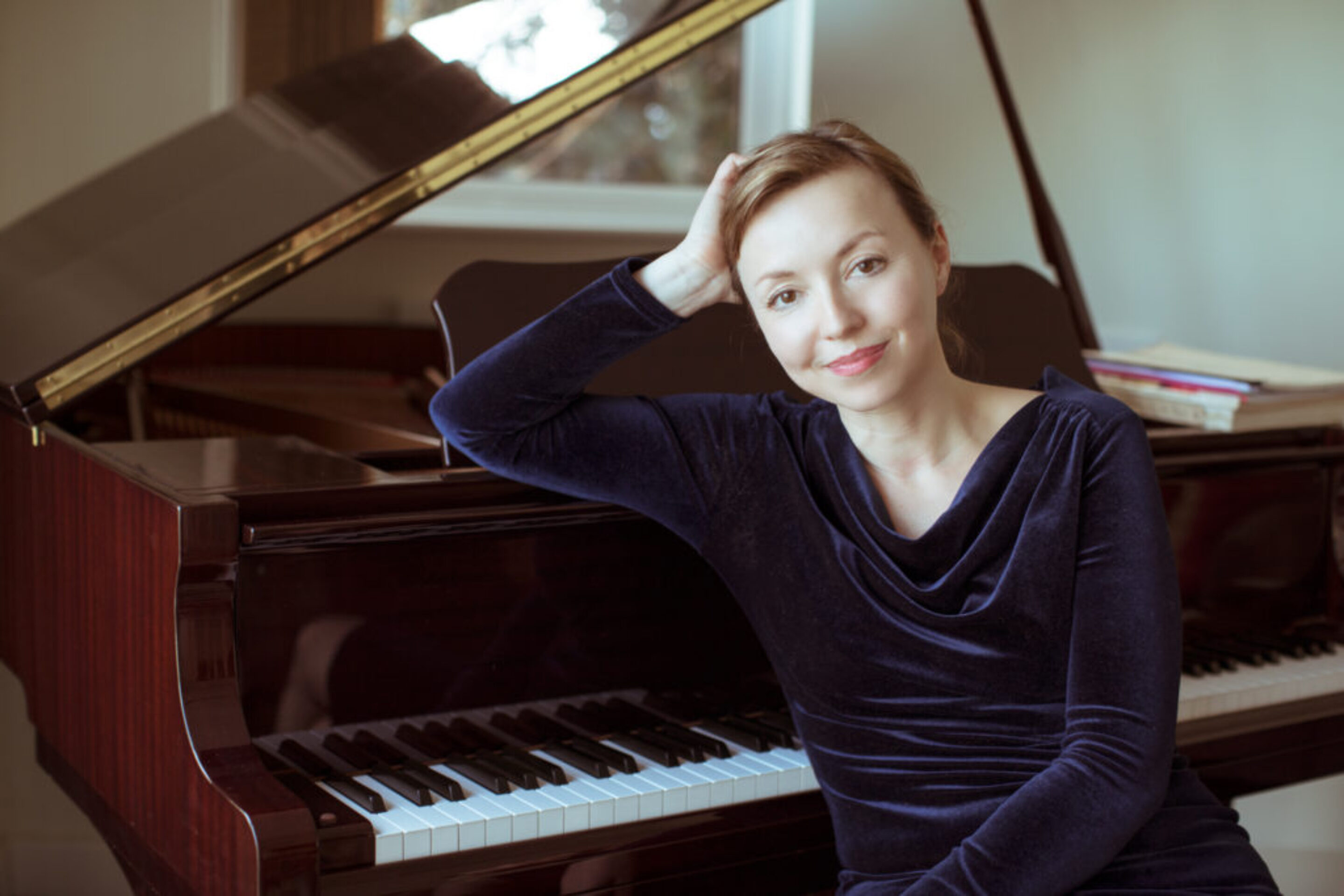 A woman in a dark blue velvet top sits beside a grand piano, resting her head on her hand and smiling gently at the camera. The piano keys and part of the open lid are visible, suggesting the world of composer Elena Langer.