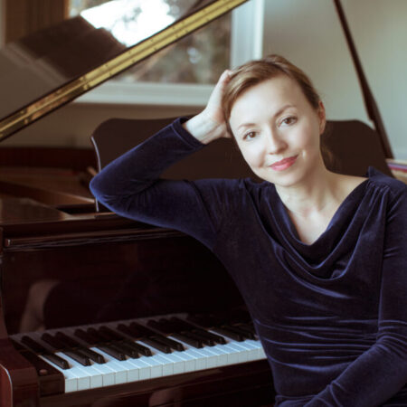 A woman in a dark blue velvet top sits beside a grand piano, resting her head on her hand and smiling gently at the camera. The piano keys and part of the open lid are visible, suggesting the world of composer Elena Langer.
