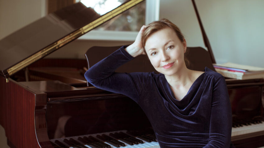 A woman in a dark blue velvet top sits beside a grand piano, resting her head on her hand and smiling gently at the camera. The piano keys and part of the open lid are visible, suggesting the world of composer Elena Langer.