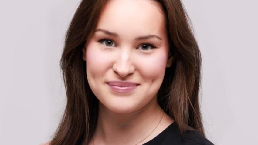 Eva Rae Martinez, a woman with long brown hair, wearing a sleeveless black top, smiles at the camera against a light grey background.