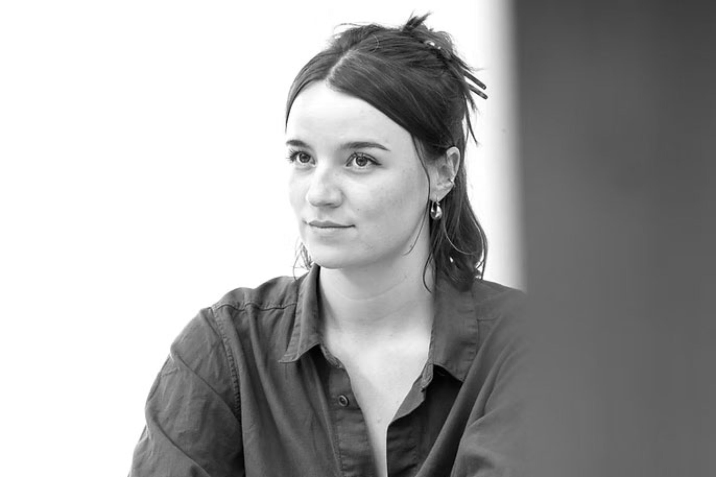 Black and white photo of a person with dark hair tied back, wearing a button-up shirt, sitting at a table with arms folded, looking slightly to the side with a neutral expression.