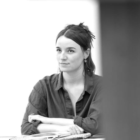 Black and white photo of a person with dark hair tied back, wearing a button-up shirt, sitting at a table with arms folded, looking slightly to the side with a neutral expression.
