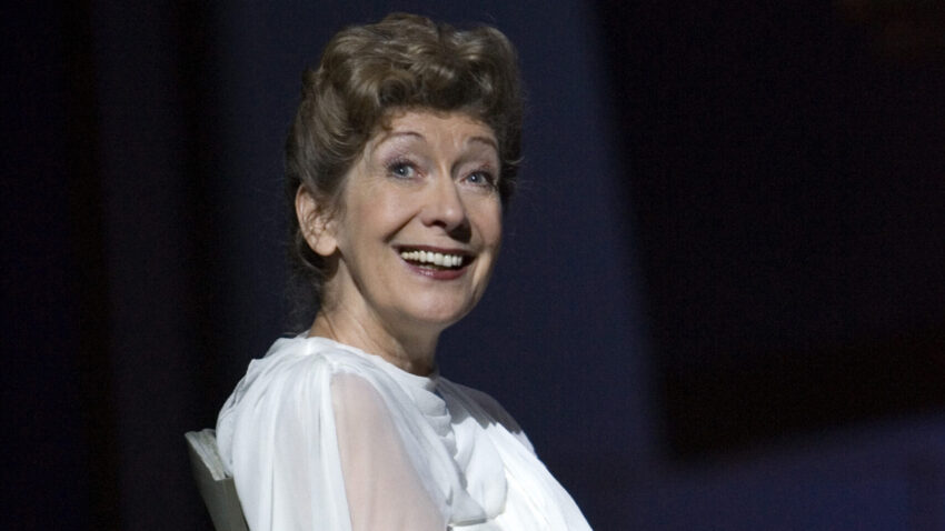 An older woman, reminiscent of celebrated soprano Dame Felicity Lott, sits on a chair in a flowing white dress, smiling warmly. She has short curly brown hair and the dark background makes her elegant ENO-inspired attire stand out.