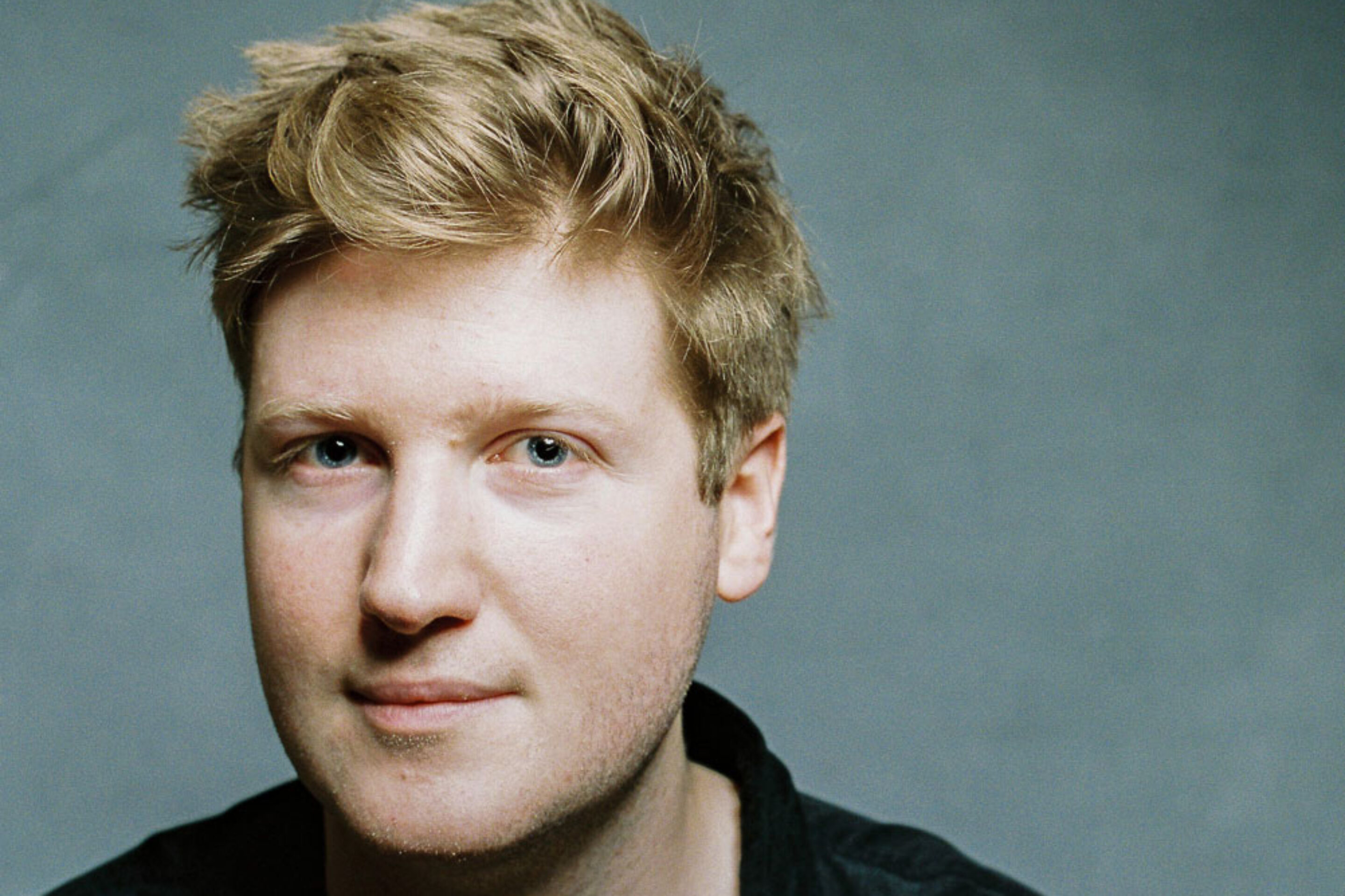 A young man with light skin and short, tousled blond hair looks at the camera with a slight smile. He is wearing a dark collared shirt and is posed against a plain, light grey background.