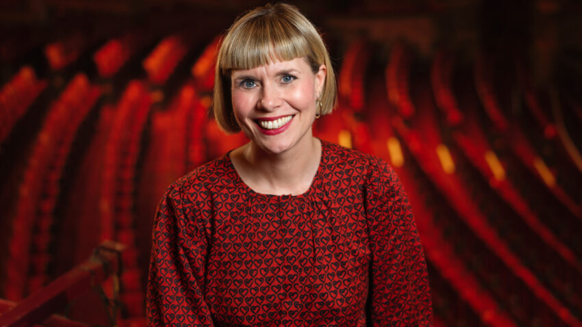Jenny, a smiling woman with short blonde hair, wearing a red patterned blouse and black trousers, stands in front of rows of red theatre seats in an ornate, warmly-lit auditorium.