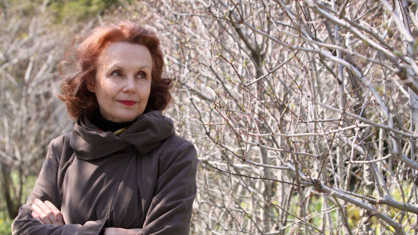 A woman with reddish hair and a brown coat stands with arms crossed, looking to the side, beside leafless trees on a bright day, evoking the introspective spirit of Kaija Saariaho.