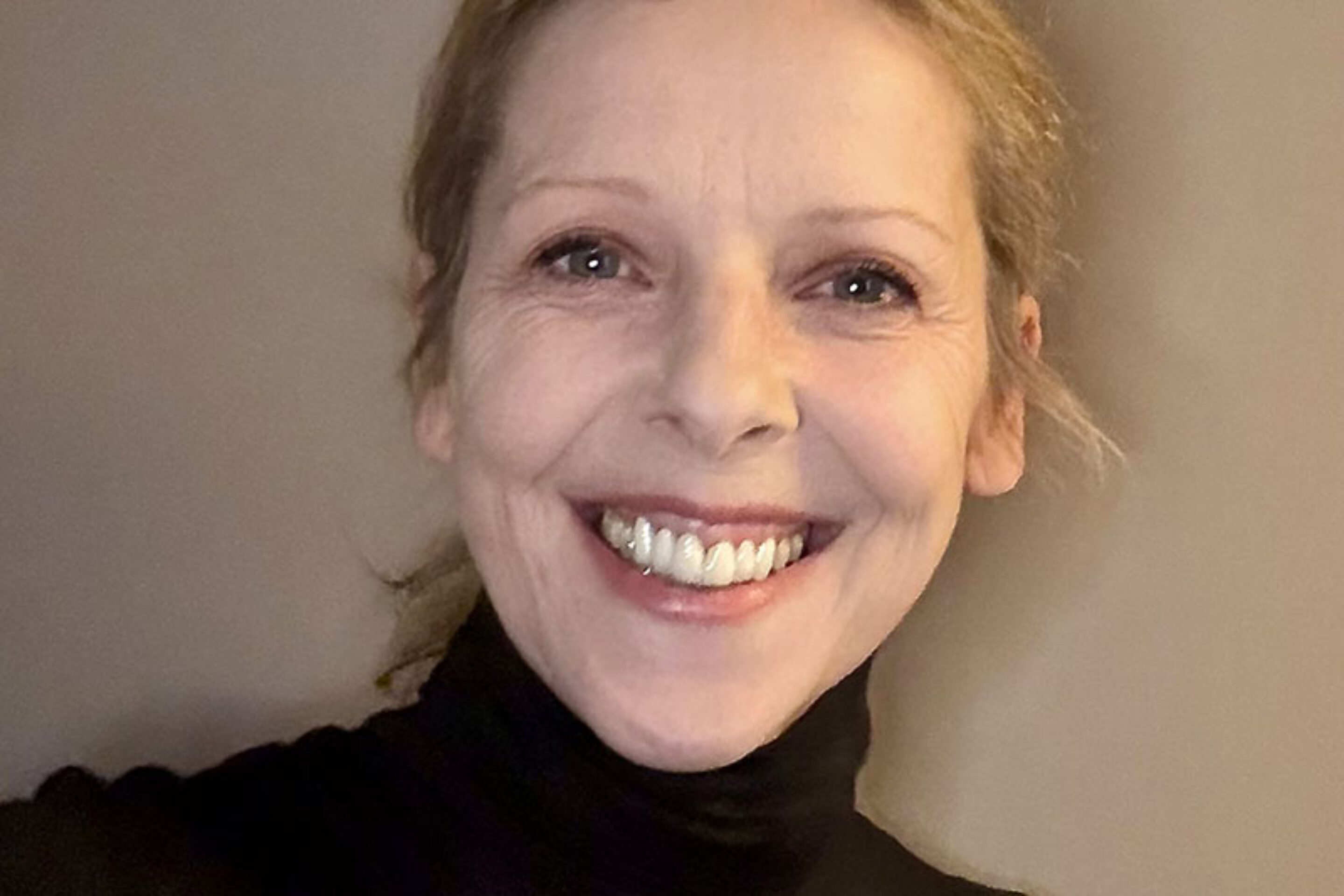 Kally Lloyd-Jones, with her blonde hair tied back, smiles brightly at the camera. She is wearing a black polo neck and is posed against a plain, neutral-coloured background.