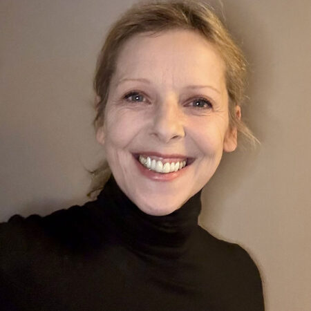 Kally Lloyd-Jones, with her blonde hair tied back, smiles brightly at the camera. She is wearing a black polo neck and is posed against a plain, neutral-coloured background.