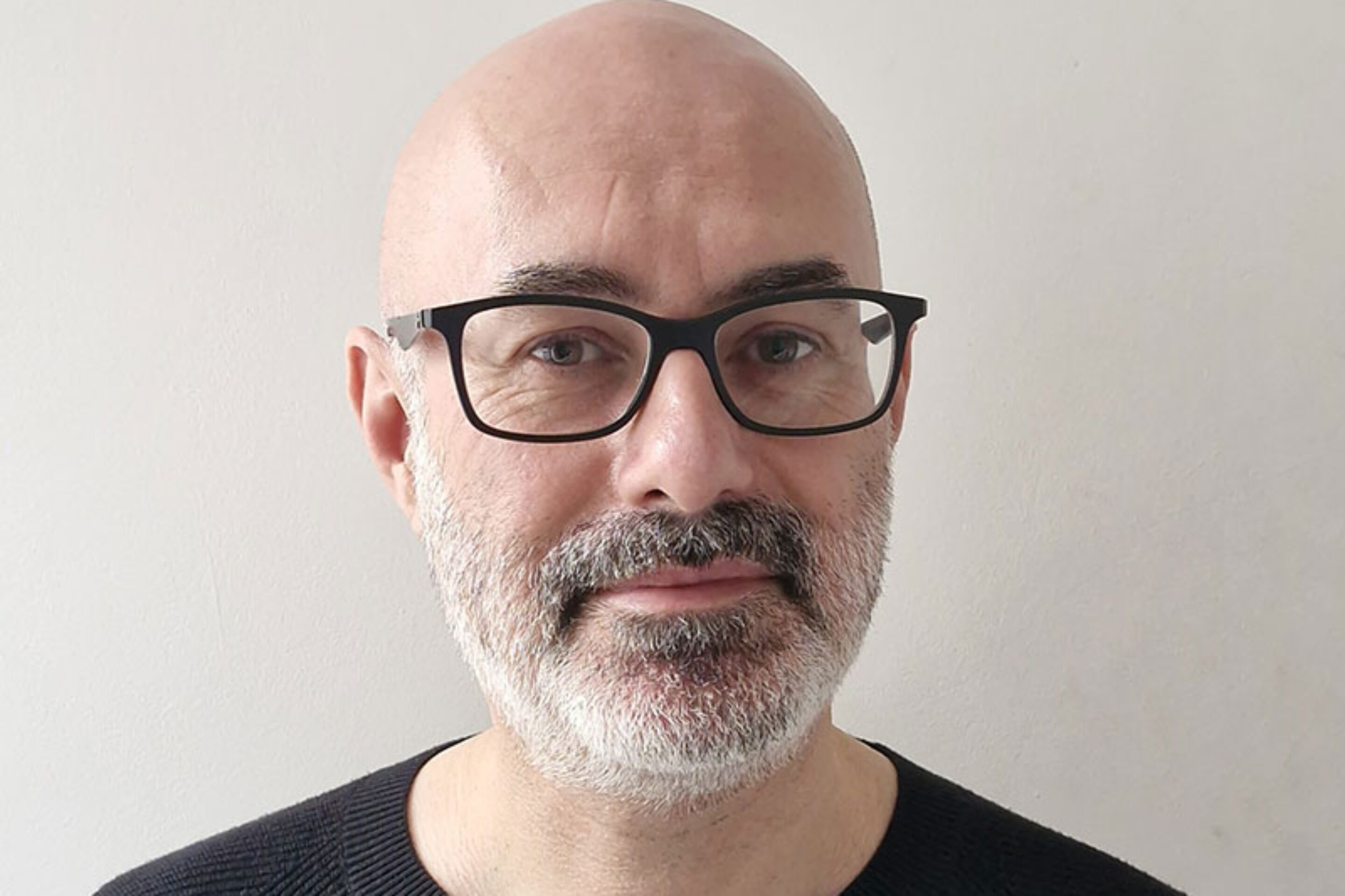 A middle-aged man with a bald head, grey beard, and black spectacles is wearing a black jumper and standing against a plain light-coloured background.