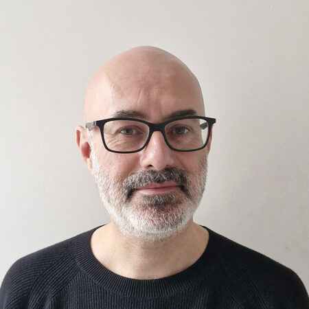 A middle-aged man with a bald head, grey beard, and black spectacles is wearing a black jumper and standing against a plain light-coloured background.
