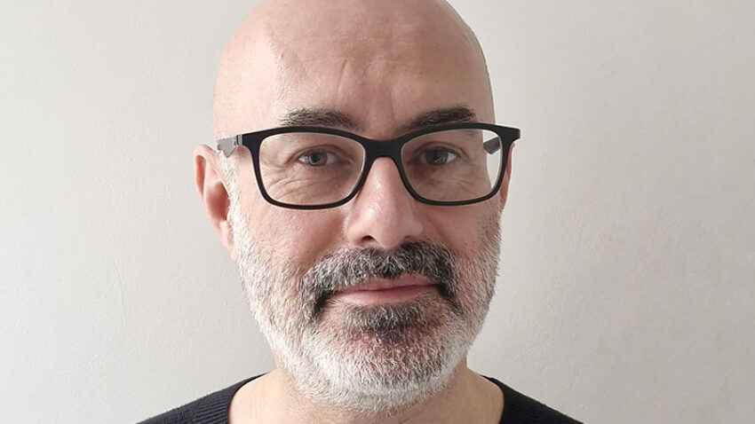 A middle-aged man with a bald head, grey beard, and black spectacles is wearing a black jumper and standing against a plain light-coloured background.