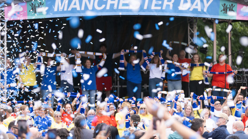 A large crowd celebrates Manchester Day with confetti falling. People in colourful shirts, including some from Salford, stand on stage under a banner that reads Manchester Day, while the audience cheers and local voices sing as photos are taken.