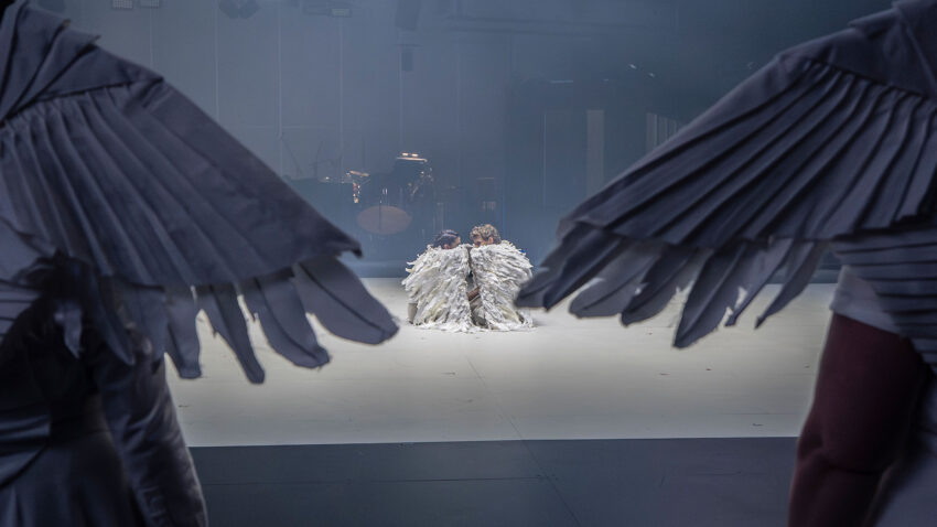 Two people wearing large white feathered wings sit together on a stage, viewed from behind two others also dressed as winged figures in the foreground, giving a dramatic, otherworldly atmosphere.