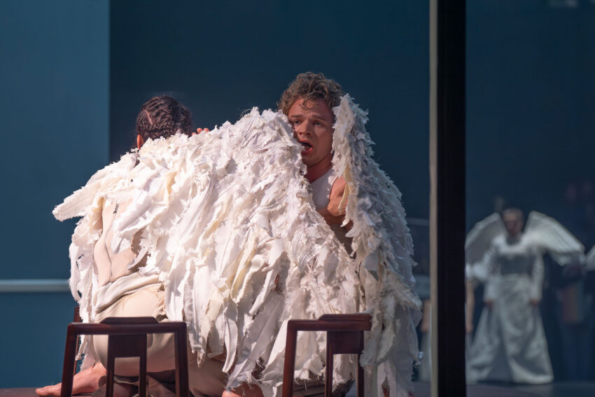 Two performers wearing white feathered costumes resembling wings sit closely facing each other on stools, whilst another person with wings is visible in the background against a blue-toned stage.