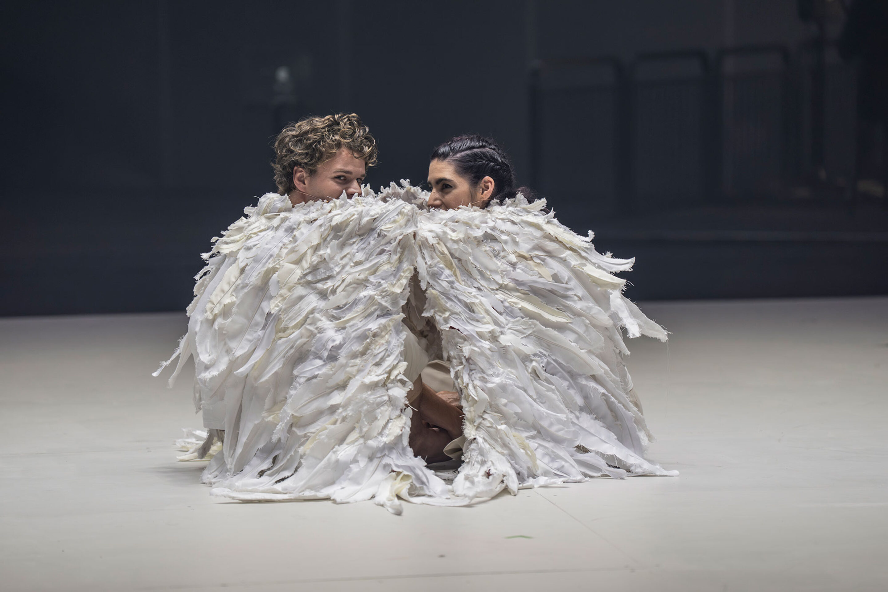 Two people crouch close together, wrapped in large, feathered, white costumes that resemble wings, on a pale, empty stage with a dark, blurred background.