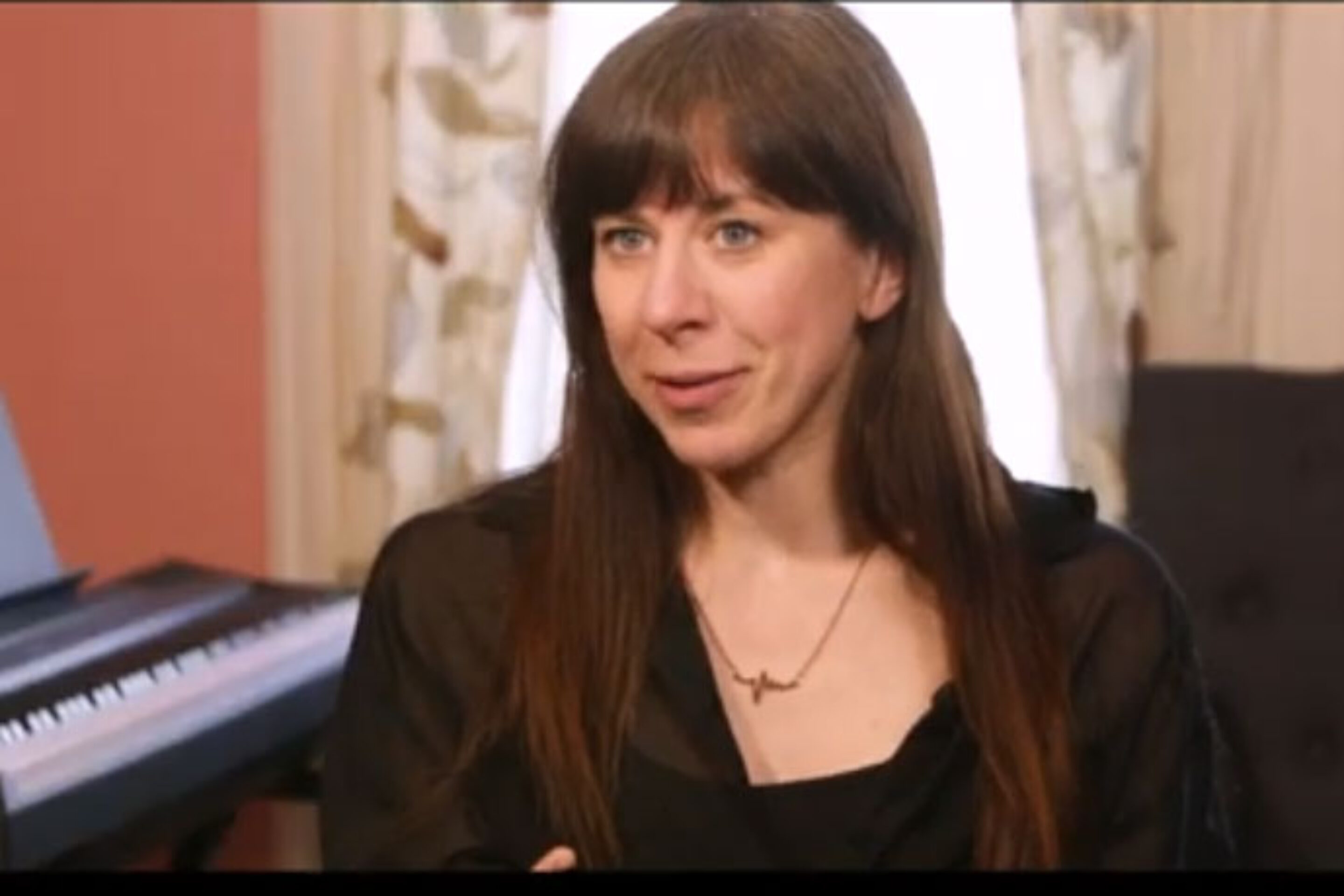 A woman with long brown hair sits indoors in front of a window with light curtains, wearing a black top and necklace. A keyboard piano is visible in the background, creating a scene reminiscent of a film introduction like Breaking the Waves.
