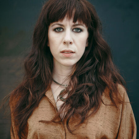 A woman with long, wavy brown hair and a fringe, wearing a brown shirt and a necklace, stands before a dark, blurred background, looking directly at the camera—reminiscent of contemporary classical composer Missy Mazzoli.