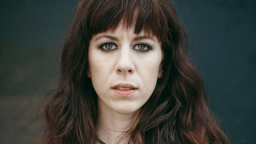A woman with long, wavy brown hair and a fringe, wearing a brown shirt and a necklace, stands before a dark, blurred background, looking directly at the camera—reminiscent of contemporary classical composer Missy Mazzoli.
