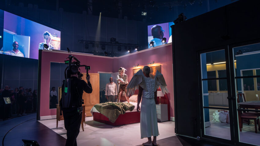 A TV set features a bedroom scene being filmed with actors, including one in angel wings and white clothing, while a camera crew records and audience members observe. Monitors display close-ups of the performers overhead.