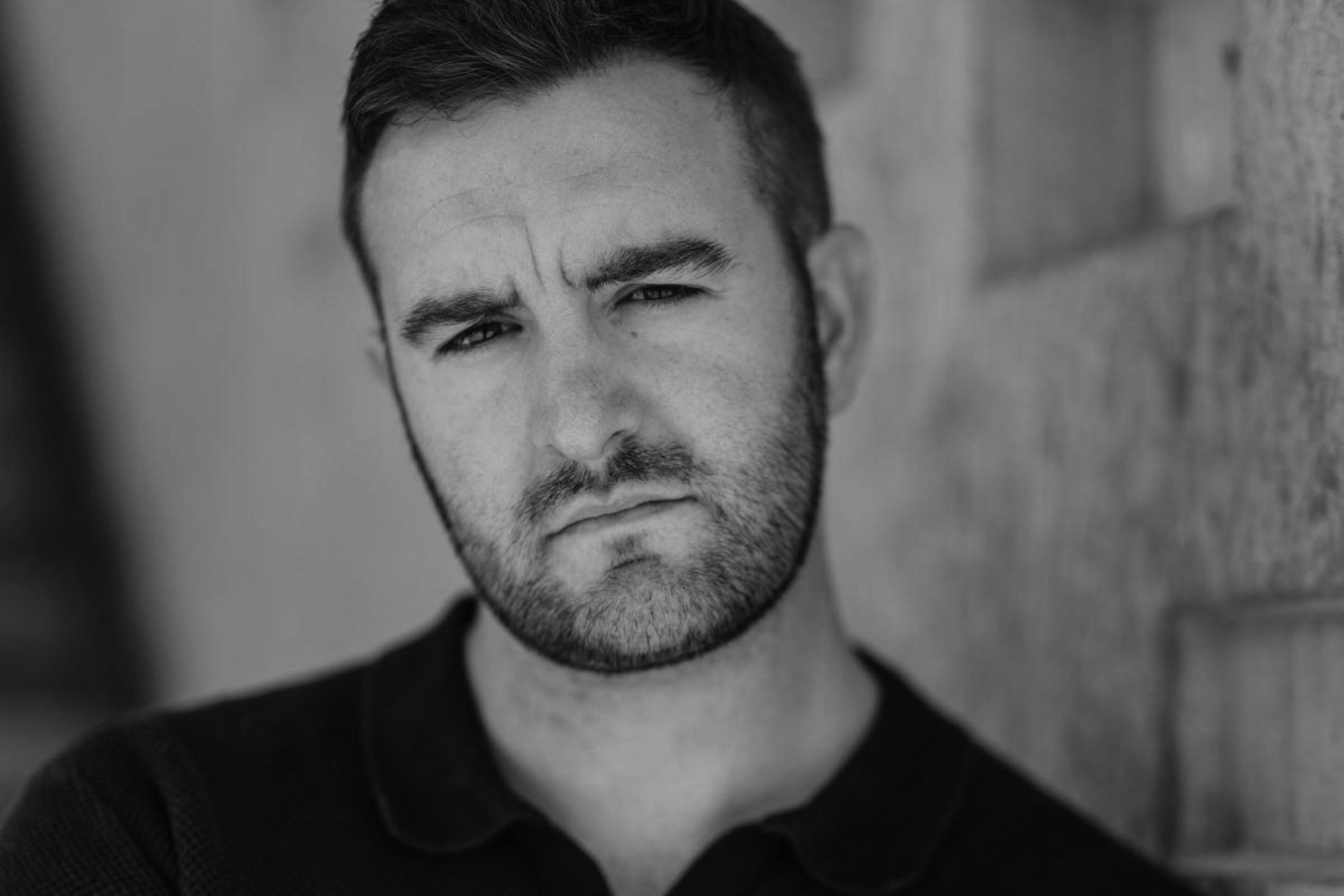 A man with short dark hair and a beard, part of the 2026/27 Harewood Artists, leans against a textured wall, looking seriously at the camera. The photo is in black and white, with a blurred background and soft lighting.