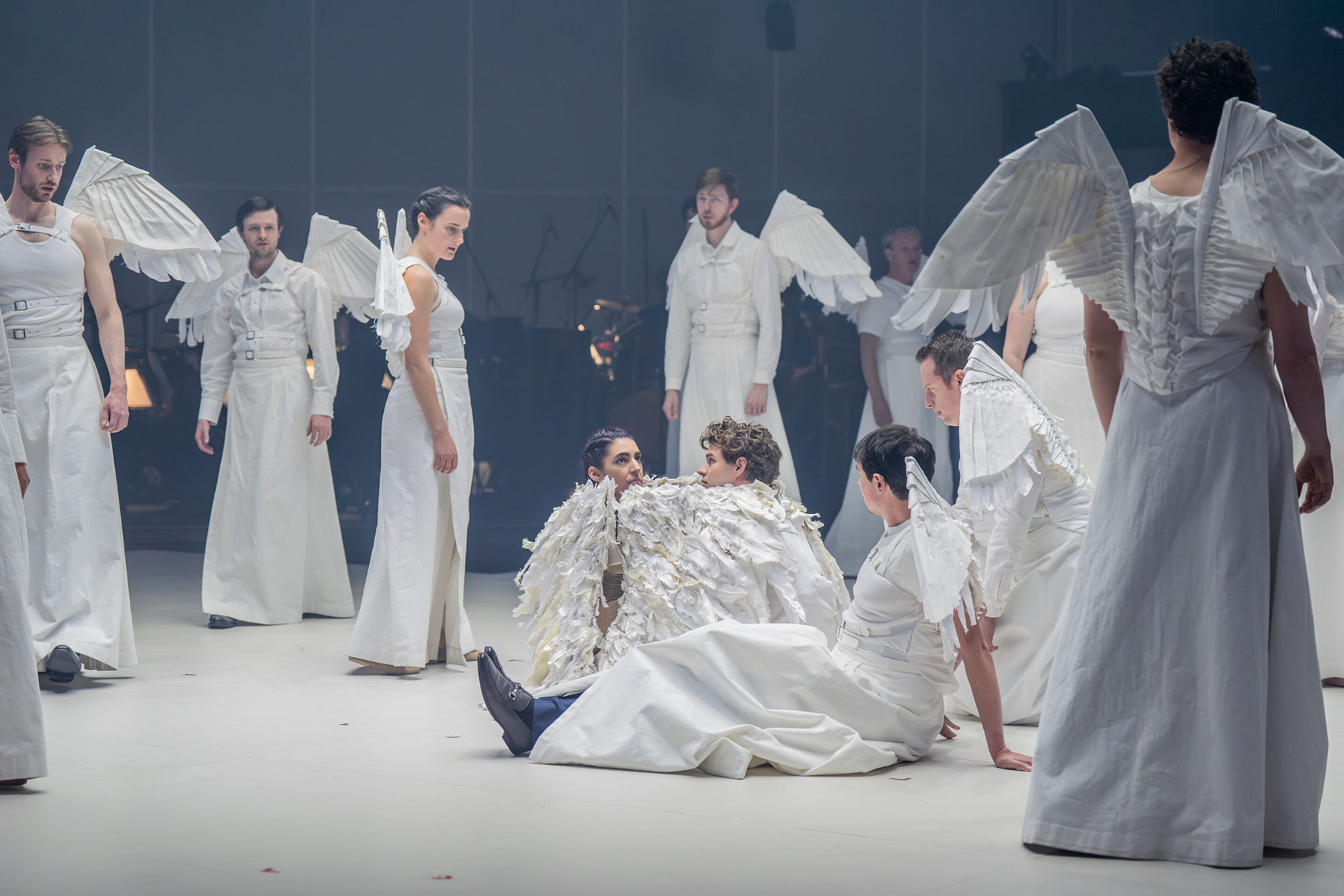 A group of performers dressed in white costumes with wing-like attachments stand and sit on a stage, some interacting closely, creating a theatrical and ethereal atmosphere.