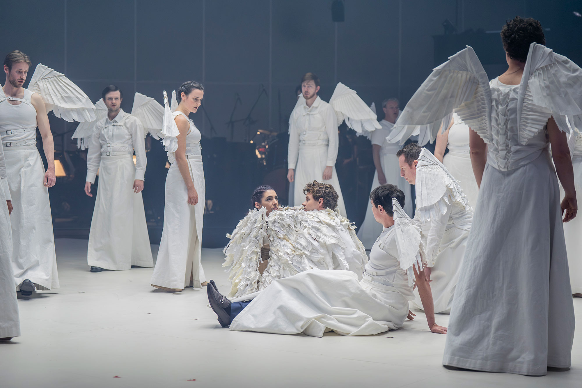 A group of performers dressed in white costumes with wing-like attachments stand and sit on a stage, some interacting closely, creating a theatrical and ethereal atmosphere.