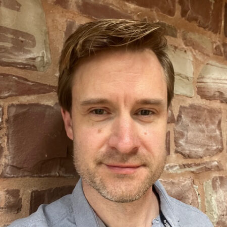 Ben Pickersgill, a man with light brown hair and light facial hair, stands in front of a stone wall wearing a light grey collared shirt, looking into the camera with a neutral expression.