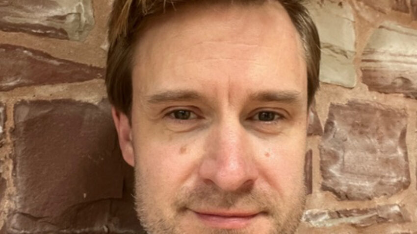 Ben Pickersgill, a man with light brown hair and light facial hair, stands in front of a stone wall wearing a light grey collared shirt, looking into the camera with a neutral expression.