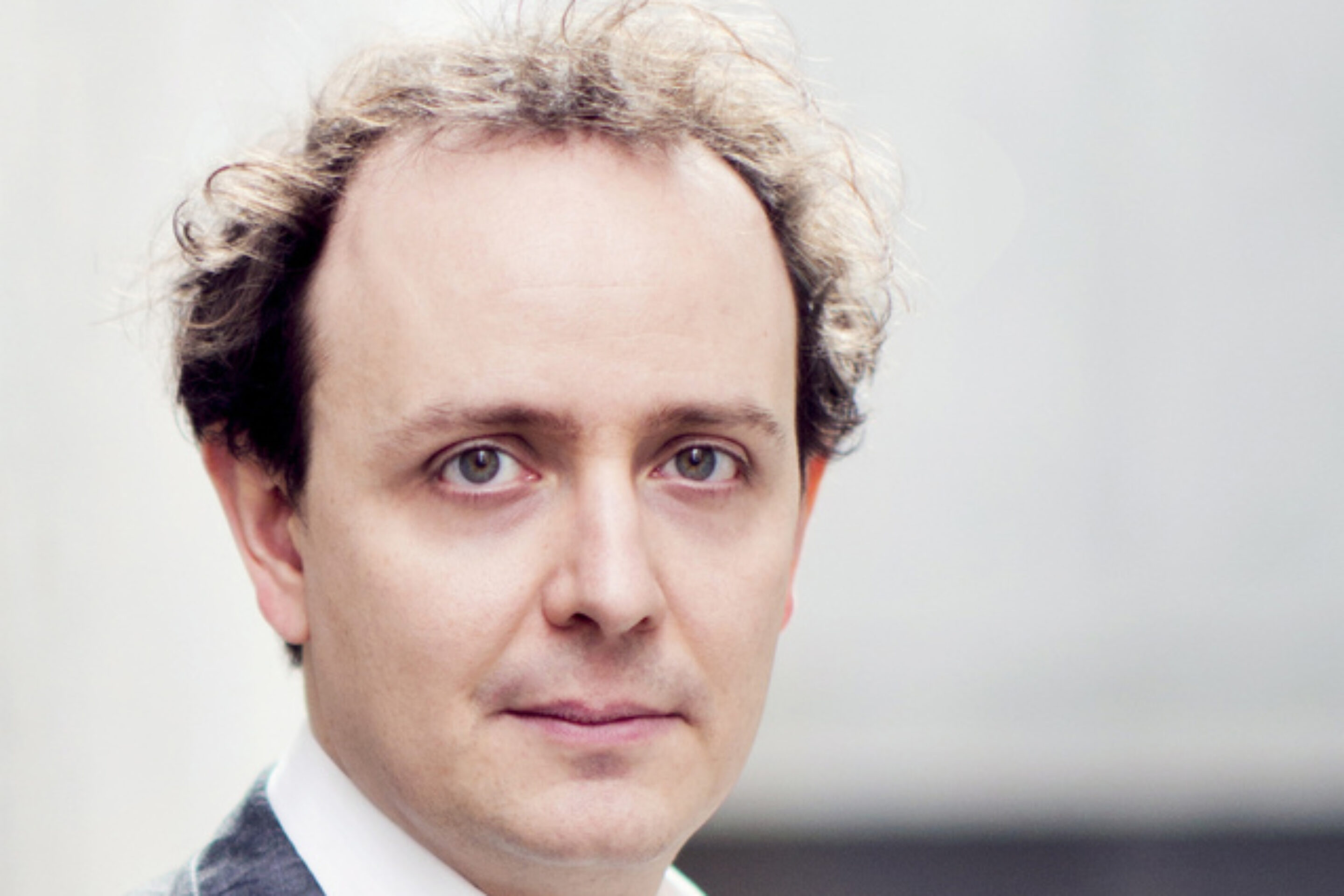 Adrian Kelly, a man with light skin and thinning, curly brown hair, is wearing a grey suit jacket and white collared shirt, looking directly at the camera with a neutral expression. The background is softly blurred.