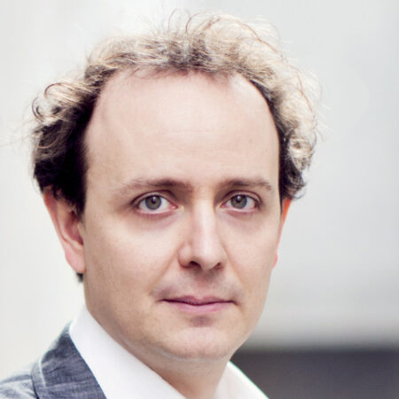 Adrian Kelly, a man with light skin and thinning, curly brown hair, is wearing a grey suit jacket and white collared shirt, looking directly at the camera with a neutral expression. The background is softly blurred.