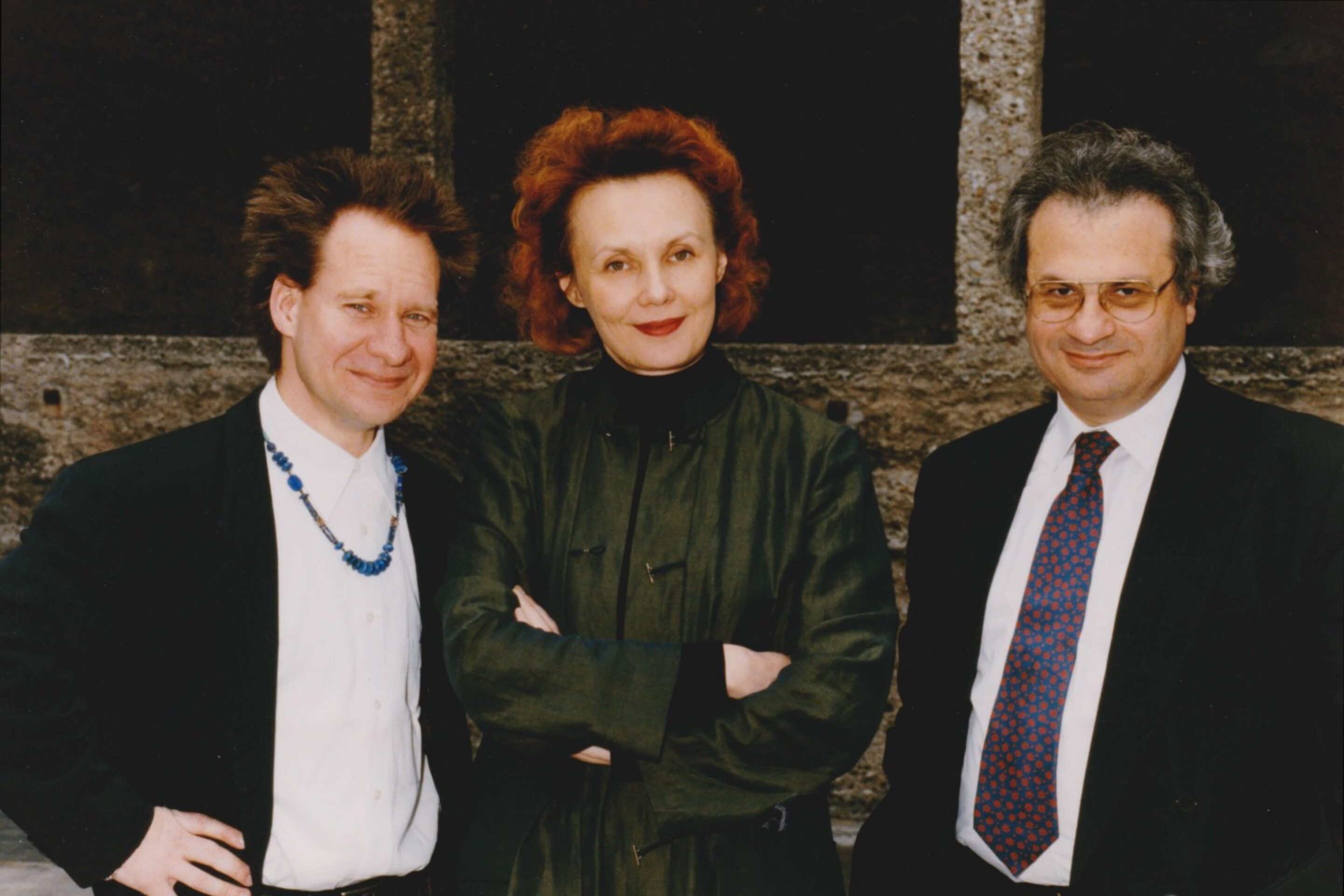 Three adults dressed in formal attire pose outdoors, evoking a scene fit for an Adriana Mater introduction—the red-haired woman stands at the centre, flanked by two men, one with a necklace and the other in a suit and tie.