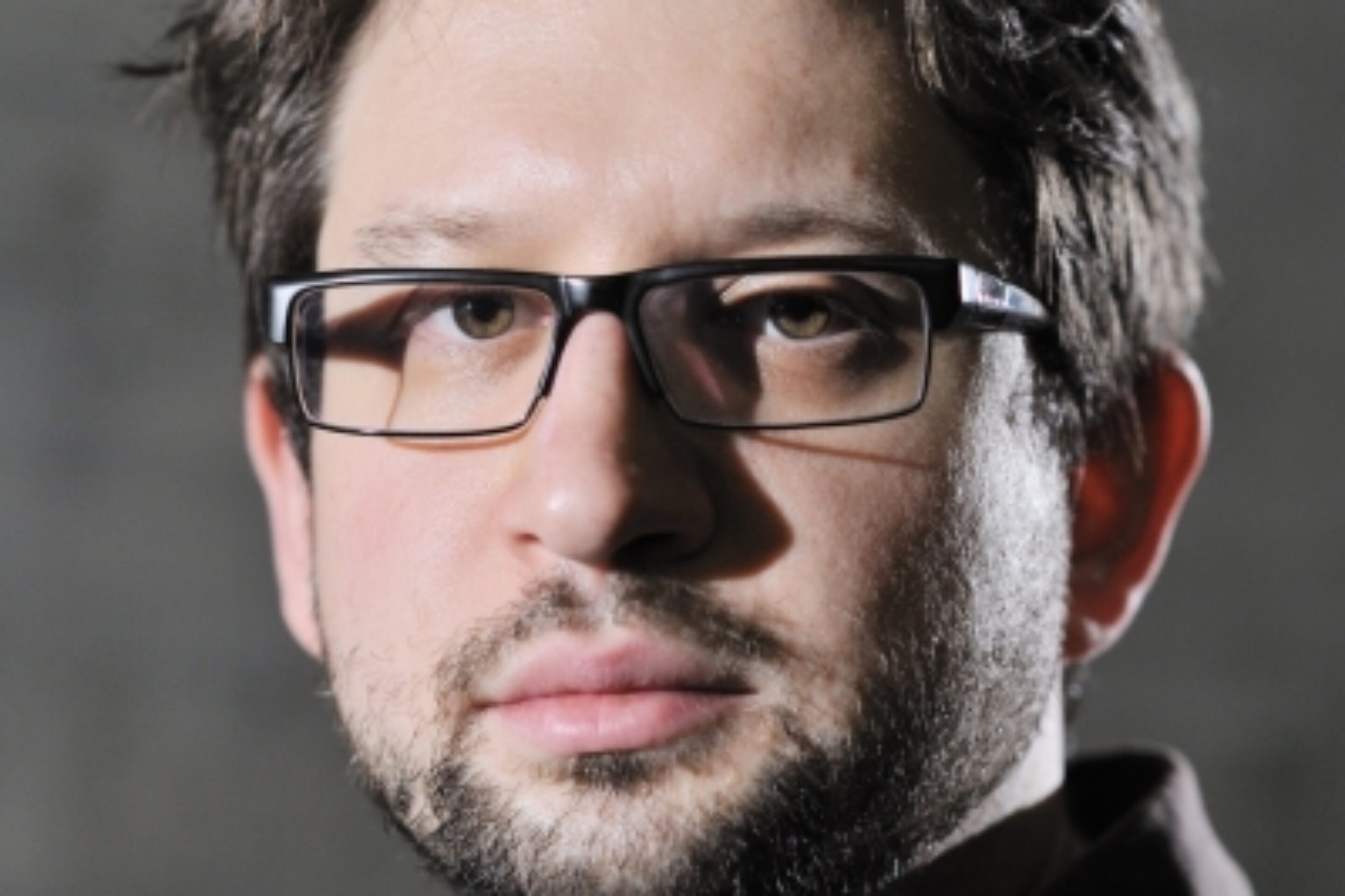 A man with short dark hair, glasses, and a trimmed beard wears a brown patterned shirt and looks directly at the camera; the background is grey and out of focus.