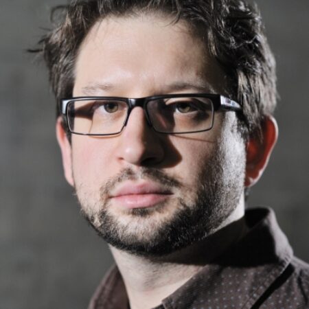 A man with short dark hair, glasses, and a trimmed beard wears a brown patterned shirt and looks directly at the camera; the background is grey and out of focus.