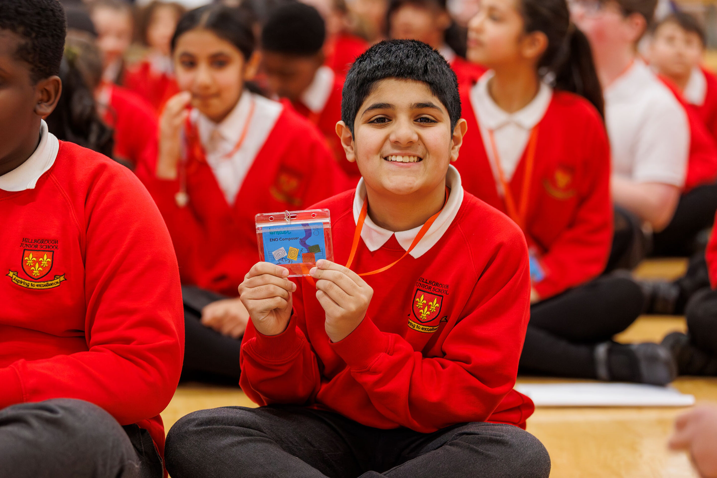 A smiling boy in a red school uniform sits cross-legged on the floor, holding up a certificate. Other pupils in matching uniforms are seated around him, also on the floor, in a group setting.