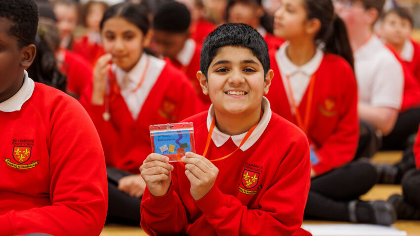 A smiling boy in a red school uniform sits cross-legged on the floor, holding up a certificate. Other pupils in matching uniforms are seated around him, also on the floor, in a group setting.