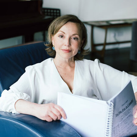Elena Langer, a woman in a white blouse sits on a blue armchair, holding an open sheet music book and looking at the camera. A piano and a table are in the background.