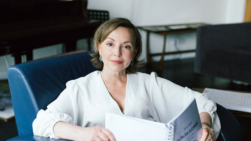 Elena Langer, a woman in a white blouse sits on a blue armchair, holding an open sheet music book and looking at the camera. A piano and a table are in the background.