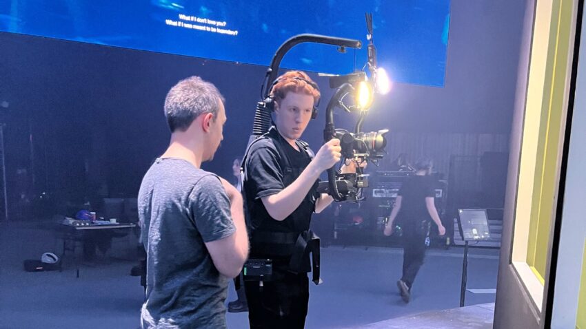 Two people stand on a stage set. One is wearing a camera rig with lights attached, preparing to film, whilst the other person speaks to him. A crew member walks in the background under blue stage lighting.