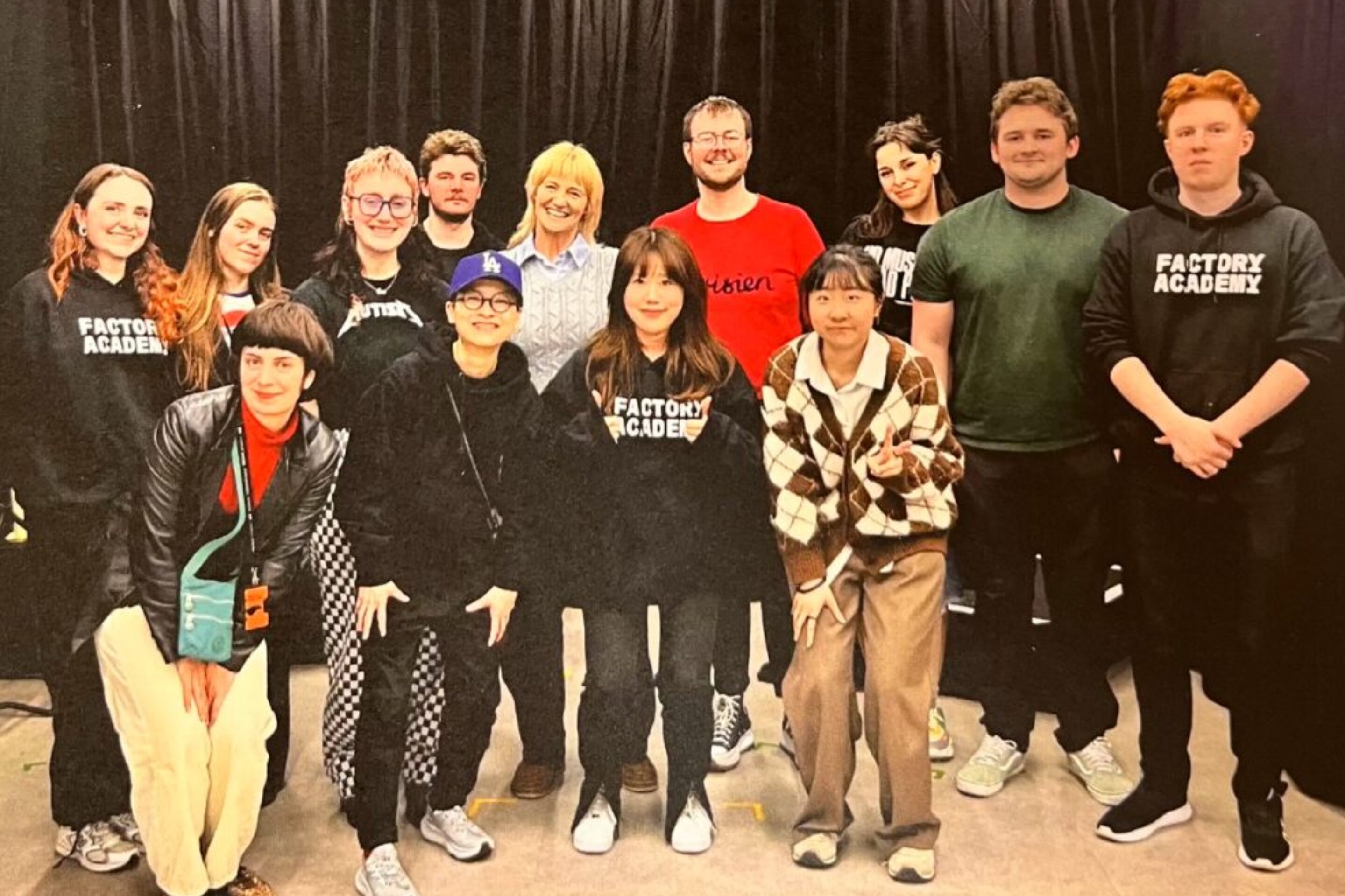 A group of thirteen people pose and smile in front of a black curtain; some wear “Factory Academy” hoodies. The words “THANK YOU!” are displayed at the bottom of the photo.