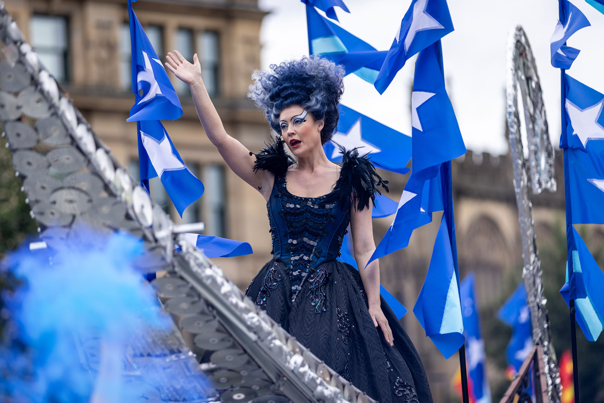 A performer with perfect pitch in an elaborate black and blue costume and dramatic make-up waves from a parade float, surrounded by blue and white flags, during a festive outdoor event.