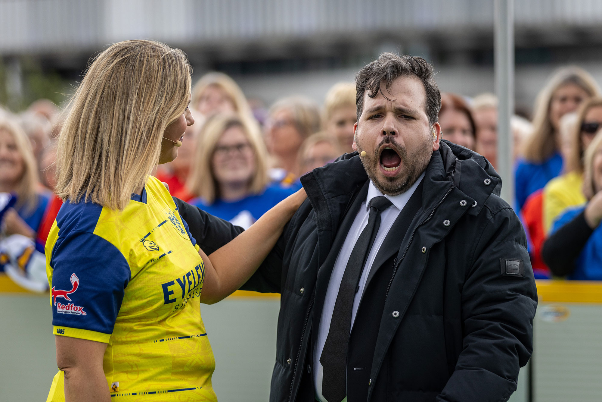 A woman in a yellow sports shirt smiles with her hand on the shoulder of a man in a black suit and tie who appears to be shouting or singing. A crowd of people in colourful shirts watches in the background.