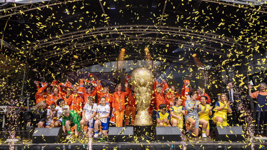 A group of people in colourful sports kits celebrate on stage with confetti falling, gathered around a large golden trophy under an arched structure.