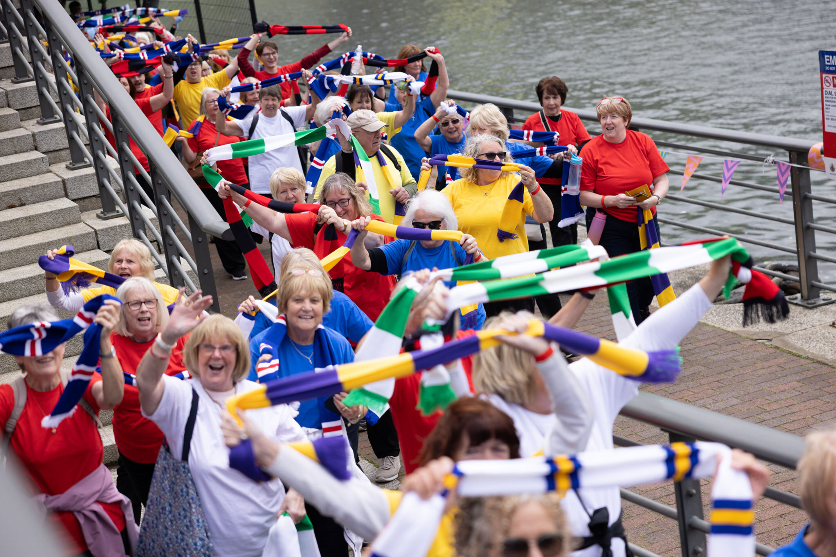 A group of smiling older adults stand by a riverside, waving colourful striped scarves and wearing bright shirts. They appear to be celebrating or participating in a joyful outdoor event.