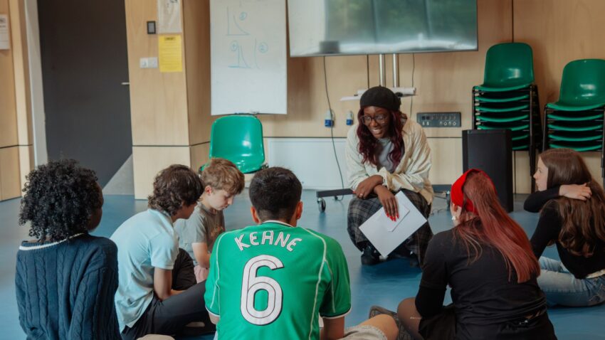A woman sits on a chair, smiling and holding papers, whilst talking to a group of six seated students in a classroom with stacked chairs, a whiteboard, and a large TV—capturing the energy of an opera pilot weekend with Greater Manchester Youth Opera and ENO.
